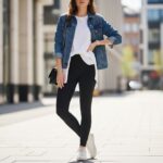 Młoda kobieta w czarnych legginsach, białej koszuli i kurtce jeansowej pozująca na chodniku w miejskim otoczeniu