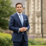 Mężczyzna w garniturze stoi pewnie na tle kościoła i zieleni w naturalnym świetle.