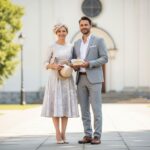 Para ubrana na chrzciny na zewnątrz, kobieta w sukience, mężczyzna w garniturze, trzymają prezent i kapelusz