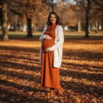 kobieta w ciąży w sukience ciążowej stoi w parku jesienią otoczona kolorowymi liśćmi